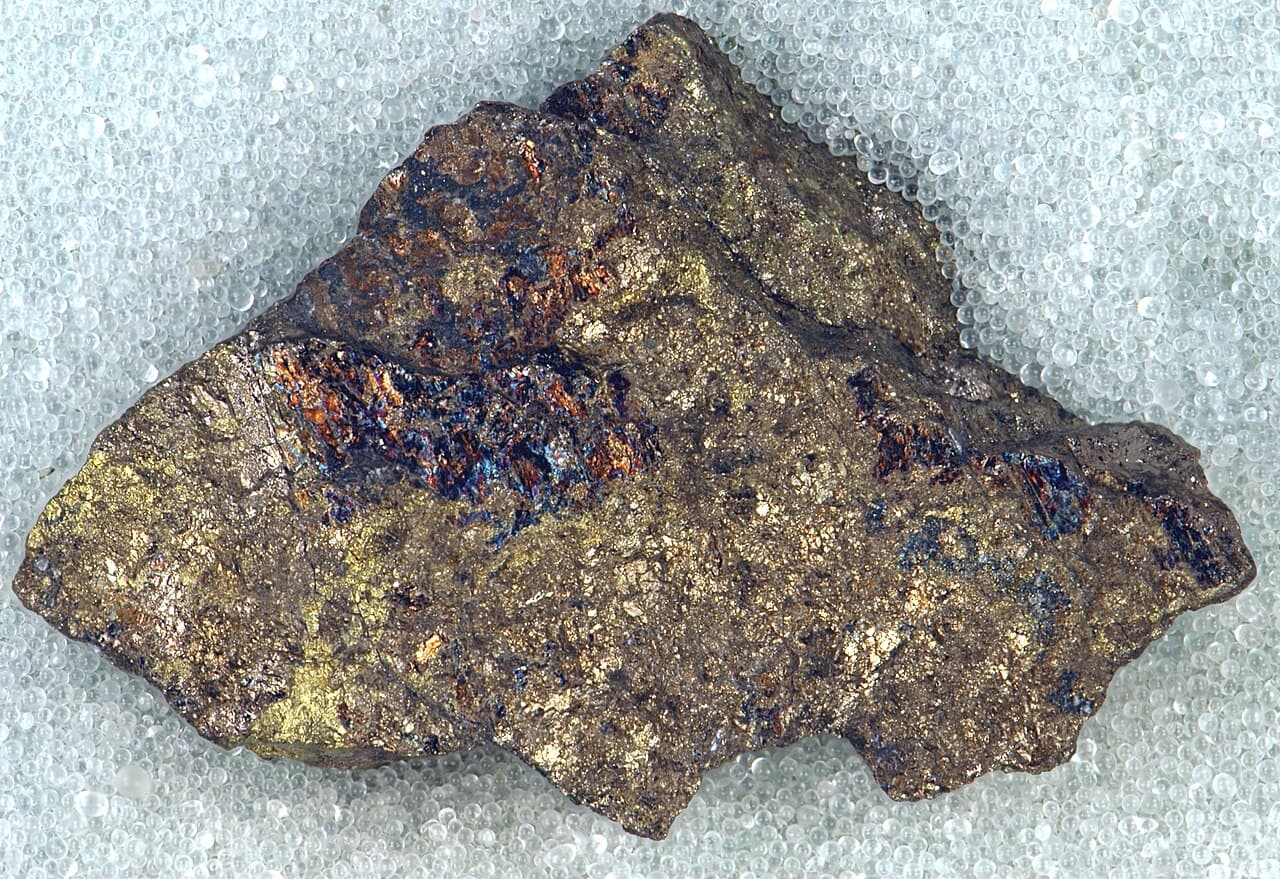 Cut slab in which bronze and purple-iridescent sulfide forms an interlocking mass through darker silicate host rock.